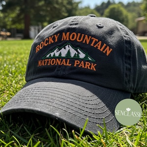 May include: A dark gray baseball cap with an embroidered logo that says "Rocky Mountain National Park" in orange and green text. The logo features a stylized mountain range with white snow peaks and green trees.