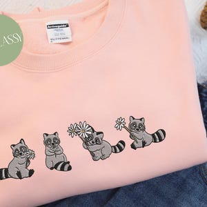 Op de afbeelding: Een roze sweatshirt met geborduurde wasberen die bloemen vasthouden. De wasberen zijn grijs met zwarte ogen en neuzen. De bloemen zijn wit met gele centra.