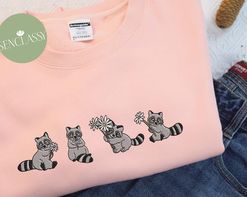 Op de afbeelding: Een roze sweatshirt met geborduurde wasberen die bloemen vasthouden. De wasberen zijn grijs met zwarte ogen en neuzen. De bloemen zijn wit met gele centra.