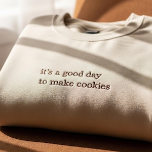 Puede incluir: Una sudadera beige doblada con la frase bordada "it's a good day to make cookies" en hilo marrón. La sudadera está hecha de un material suave y está doblada cuidadosamente sobre una superficie marrón.