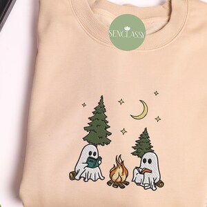 May include: A beige sweatshirt with an embroidered design of two ghosts sitting by a campfire in the woods. The ghosts are holding books and there are stars and a crescent moon in the sky. The text "Senclassy" is embroidered on the top of the sweatshirt.