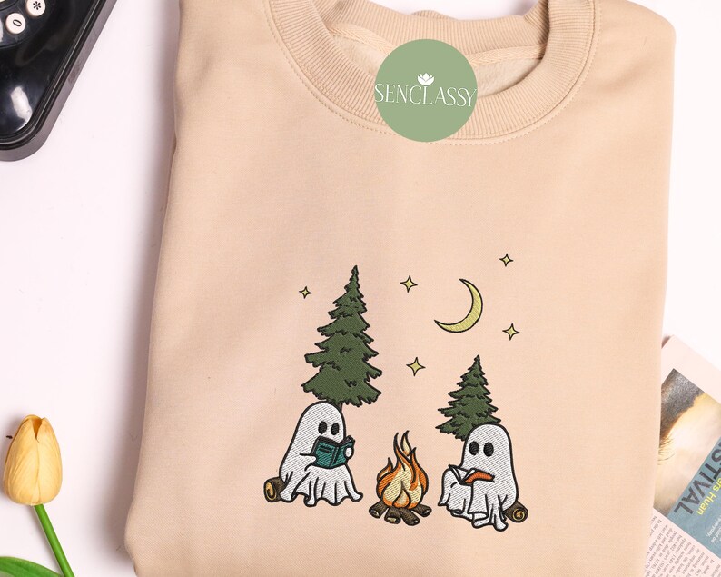 May include: A beige sweatshirt with an embroidered design of two ghosts sitting by a campfire in the woods. The ghosts are holding books and there are stars and a crescent moon in the sky. The text "Senclassy" is embroidered on the top of the sweatshirt.