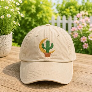 Könnte beinhalten: Eine beige Baseballkappe mit einem gestickten Motiv auf der Vorderseite. Die Stickerei zeigt einen grünen Kaktus in einem braunen Topf vor einem gelben Halbmond. Die Kappe hat einen gebogenen Schirm und eine strukturierte sechsteilige Krone.
