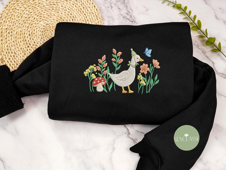 May include: Black sweatshirt with a white goose embroidered on the front. The goose is wearing a small hat and is surrounded by flowers and a mushroom.
