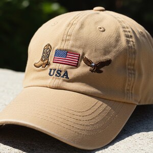 Może przedstawiać: Beżowa czapka z daszkiem z haftowaną flagą Stanów Zjednoczonych, kowbojskim butem i orłem. Na czapce jest również wyszyte słowo "USA".