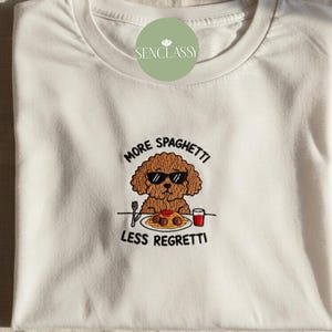 May include: White t-shirt with an embroidered design featuring a brown poodle wearing sunglasses, eating spaghetti. The text "MORE SPAGHETTI LESS REGRETTI" is above and below the dog. The shirt is folded.