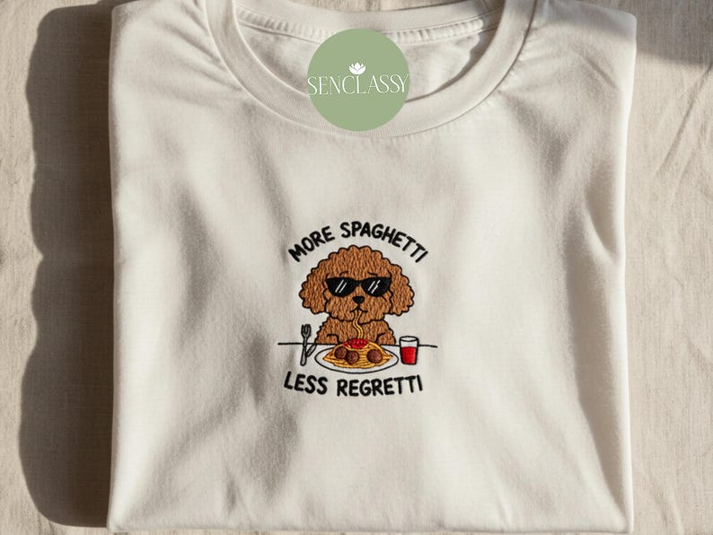 May include: White t-shirt with an embroidered design featuring a brown poodle wearing sunglasses, eating spaghetti. The text "MORE SPAGHETTI LESS REGRETTI" is above and below the dog. The shirt is folded.