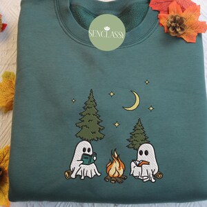 May include: A teal green sweatshirt with an embroidered design of two ghosts reading books by a campfire in the woods. The design includes trees, stars, and a crescent moon. The text "Senclassy" is embroidered on the front of the sweatshirt.