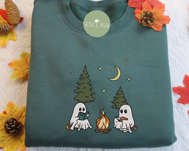 May include: A teal green sweatshirt with an embroidered design of two ghosts reading books by a campfire in the woods. The design includes trees, stars, and a crescent moon. The text "Senclassy" is embroidered on the front of the sweatshirt.