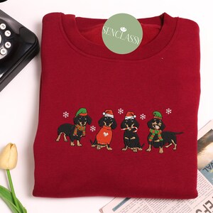 May include: A burgundy sweatshirt featuring an embroidered design of four dachshunds dressed in festive holiday attire. Each dog wears a hat, scarf, or sweater, with snowflakes scattered around them. The sweatshirt has a crew neck.