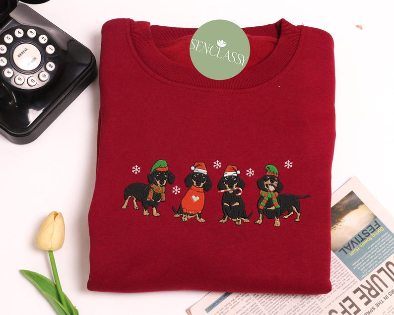 May include: A burgundy sweatshirt featuring an embroidered design of four dachshunds dressed in festive holiday attire. Each dog wears a hat, scarf, or sweater, with snowflakes scattered around them. The sweatshirt has a crew neck.