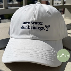 Puede incluir: Gorra de béisbol blanca con el texto bordado en azul marino "save water drink margs.". La gorra tiene una visera curva y un gráfico de copa de margarita. El sombrero está hecho de una tela suave y texturizada.