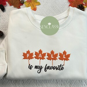 May include: White sweatshirt with the text "FALL is my favorite" embroidered in orange and black. The word "FALL" is designed with orange maple leaves. The sweatshirt has a round neckline and long sleeves. Fall-themed apparel.