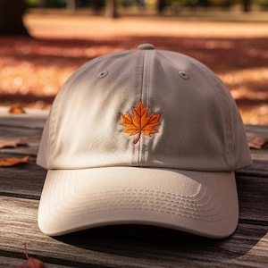 Puede incluir: Gorra de béisbol beige con visera curva. Una hoja de arce naranja está bordada en la parte delantera. El sombrero está sobre una superficie de madera con hojas caídas en el fondo.