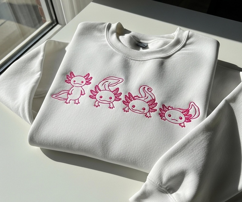 May include: White sweatshirt featuring a pink embroidered design of four axolotls. The axolotls are in various poses, with detailed gills and smiling faces. The sweatshirt is made of a soft material and has a crew neck.