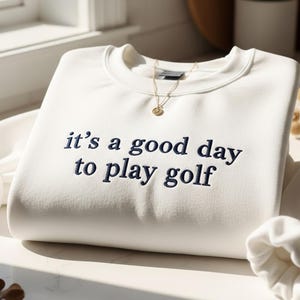 Pu&ograve; includere: Felpa color crema con la frase ricamata "it's a good day to play golf" in blu navy. Una collana d'oro con un piccolo ciondolo &egrave; appoggiata sulla camicia. Le maniche sono piegate e la camicia &egrave; ben riposta.