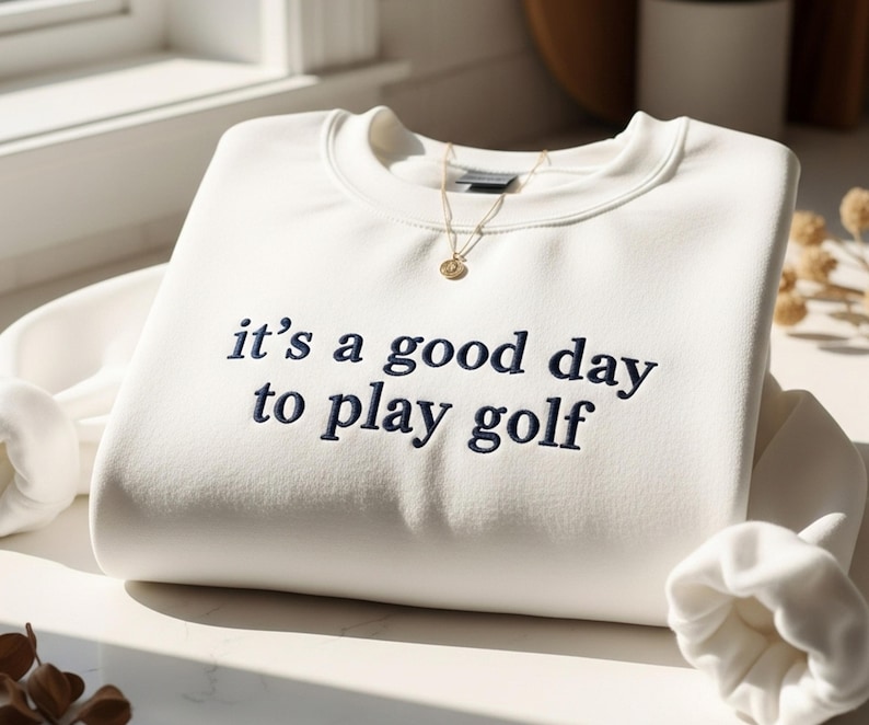 Pu&ograve; includere: Felpa color crema con la frase ricamata "it's a good day to play golf" in blu navy. Una collana d'oro con un piccolo ciondolo &egrave; appoggiata sulla camicia. Le maniche sono piegate e la camicia &egrave; ben riposta.