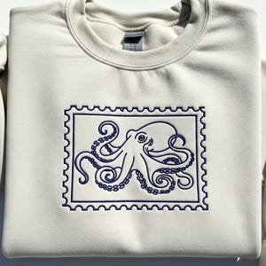 May include: A cream-colored sweatshirt featuring a navy blue embroidered octopus design within a postage stamp frame. The octopus has detailed tentacles and a smiling face. The sweatshirt is folded, showing the crew neck and a portion of the sleeve.