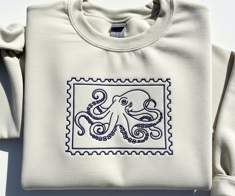 May include: A cream-colored sweatshirt featuring a navy blue embroidered octopus design within a postage stamp frame. The octopus has detailed tentacles and a smiling face. The sweatshirt is folded, showing the crew neck and a portion of the sleeve.