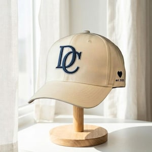 May include: A cream-colored baseball cap with a curved brim, featuring a navy blue embroidered "DC" logo on the front. A small black heart and the text "est. 2025" are embroidered on the side. The cap is displayed on a wooden stand.