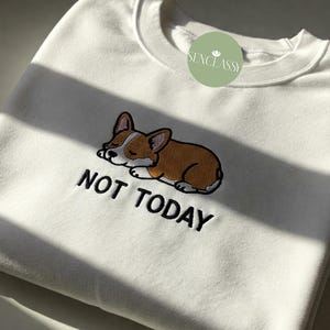 以下が含まれることがあります： 白いスウェットシャツに、寝ているコーギーの漫画と「NOT TODAY」という刺繍が施されています。