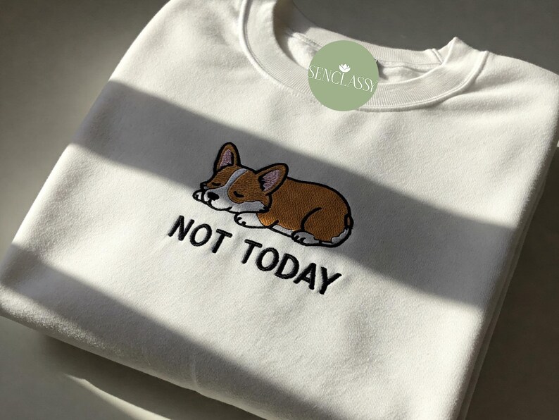 以下が含まれることがあります： 白いスウェットシャツに、寝ているコーギーの漫画と「NOT TODAY」という刺繍が施されています。