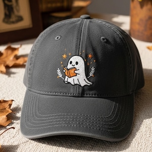 Puede incluir: Gorra de béisbol gris con un fantasma bordado leyendo un libro. El fantasma es blanco con una cara negra, sosteniendo un libro naranja. Estrellas naranjas y ramitas de hojas rodean al fantasma. La gorra tiene una visera curva y una correa de tela.