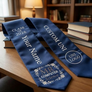 以下が含まれることがあります： 白の刺繍テキストが入ったネイビーブルーの卒業用サッシュ。「CLASS OF 2026」、「Your Name」、「CUSTOM UNI」、「MR COMPUTER Science」と書かれています。大学のロゴも表示されています。サッシュは本の上にかけられています。