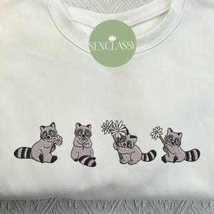 Op de afbeelding: Witte trui met geborduurde wasberen die bloemen vasthouden. De wasberen zijn grijs met zwarte ogen en neuzen. De bloemen zijn wit met gele centra.