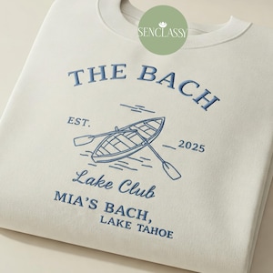 May include: A white sweatshirt with blue embroidered text that reads "The Bach, Est. 2025, Lake Club, Mia's Bach, Lake Tahoe". The design also includes a blue embroidered illustration of a boat with oars.