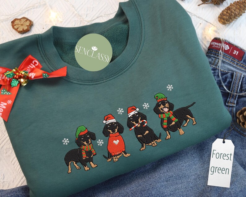 May include: Forest green sweatshirt with embroidered dachshunds wearing Christmas hats and scarves. The dogs are black and tan, with snowflakes around them. The sweatshirt has a crew neck and long sleeves.