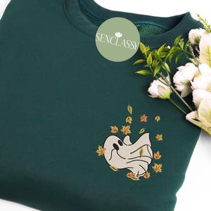 Puede incluir: Una sudadera verde oscuro con un diseño bordado de un fantasma con hojas que caen. El diseño está en tonos amarillos y naranjas.