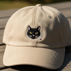 May include: A tan baseball cap with a black cat face embroidered on the front. The cat has yellow eyes and a cigarette in its mouth. The cap is made of a soft material and has a curved brim.