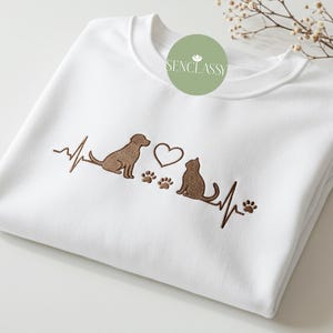 Op de afbeelding: Witte trui met een bruin geborduurd ontwerp van een hond en een kat met een hart en poot afdrukken. Het ontwerp bevat een hartslaglijn.