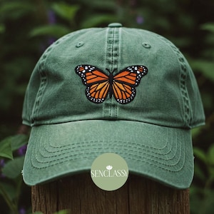 Puede incluir: Una gorra de béisbol verde con un diseño de mariposa monarca bordada. La mariposa es naranja, negra y blanca. La gorra tiene la palabra "Senclassy" bordada en la parte delantera.