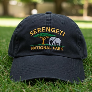May include: A black baseball cap with embroidered text and a graphic. The text reads "SERENGETI NATIONAL PARK" in gold and white. The graphic features a tree and an elephant. The cap is sitting on green grass.