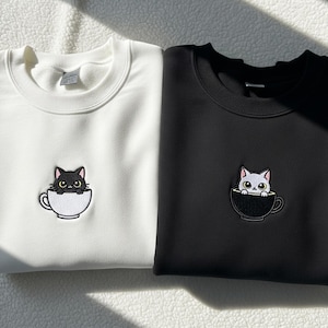Op de afbeelding: Twee sweatshirts, één witte en één zwarte, elk met een geborduurd kattenontwerp. De kat zit in een theekopje. Het witte sweatshirt heeft een zwarte kat en het zwarte sweatshirt heeft een witte kat.