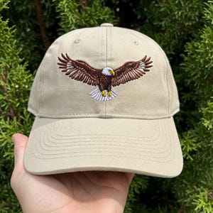 May include: A beige baseball cap with an embroidered image of a bald eagle with brown and white feathers, spreading its wings.