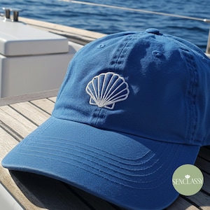 Puede incluir: Una gorra de béisbol azul con un diseño de concha bordado en blanco. La gorra tiene una visera curvada y un logotipo blanco en el lateral que dice "Senclassi".