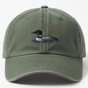 Puede incluir: Gorra de béisbol verde oliva con un diseño bordado de un somorgujo nadando en el agua. El somorgujo tiene la cabeza negra, el cuerpo moteado blanco y negro y el ojo rojo. La gorra tiene una visera curva y una correa ajustable.