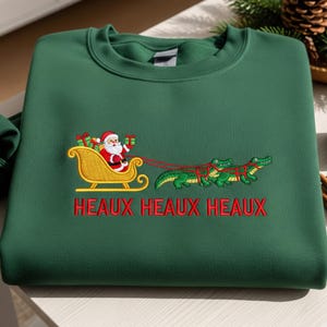 May include: A dark green sweatshirt featuring an embroidered design of Santa Claus in a gold sleigh pulled by alligators. The text "HEAUX HEAUX HEAUX" is embroidered in red below the design. A festive holiday-themed sweatshirt.