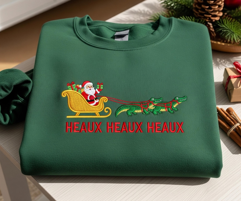 May include: A dark green sweatshirt featuring an embroidered design of Santa Claus in a gold sleigh pulled by alligators. The text "HEAUX HEAUX HEAUX" is embroidered in red below the design. A festive holiday-themed sweatshirt.