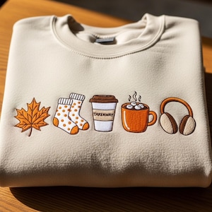 May include: Cream-coloured sweatshirt with embroidered autumn motifs. Includes a maple leaf, socks with orange spots, a takeaway coffee cup, a mug of hot chocolate with marshmallows, and headphones. The coffee cup has the word "TAKEAWAY".