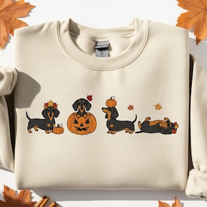 May include: Cream-colored sweatshirt featuring a dachshund dog design with fall-themed elements. The design includes dachshunds with pumpkins, leaves, and a pumpkin-shaped jack-o'-lantern. The sweatshirt has long sleeves and a crew neck.