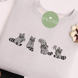 Op de afbeelding: Een grijs sweatshirt met een geborduurd ontwerp van vier wasberen die bloemen vasthouden. De wasberen kijken allemaal naar voren en hebben zwarte ogen en neuzen. De bloemen zijn wit en hebben vijf bloemblaadjes. De tekst "Senclassy" is in het groen geborduurd in de linkerbovenhoek van het sweatshirt.