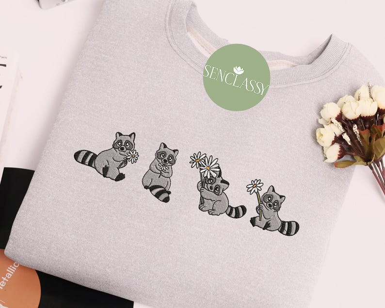 Op de afbeelding: Een grijs sweatshirt met een geborduurd ontwerp van vier wasberen die bloemen vasthouden. De wasberen kijken allemaal naar voren en hebben zwarte ogen en neuzen. De bloemen zijn wit en hebben vijf bloemblaadjes. De tekst "Senclassy" is in het groen geborduurd in de linkerbovenhoek van het sweatshirt.