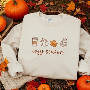 May include: Cream-colored sweatshirt with a fall-themed design. The design includes a coffee cup, pumpkin, leaf, and beanie, with the words "cozy season" in brown script. The sweatshirt is surrounded by pumpkins and fall foliage.