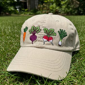 Puede incluir: Una gorra de béisbol beige con verduras bordadas. Las verduras incluyen una zanahoria, una remolacha, dos rábanos y una cebolla verde.