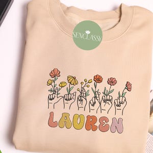 May include: A beige sweatshirt with a floral design and the name "LAUREN" in colorful, retro-style lettering.