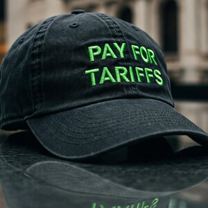 May include: A black baseball cap with a green embroidered message that reads "PAY FOR TARIFFS".